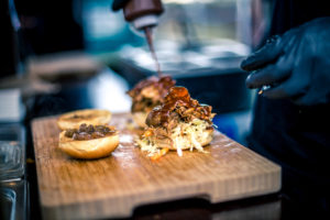 Jemand schüttet Soße auf einen Hamburger-Patty auf einem Holzbrett